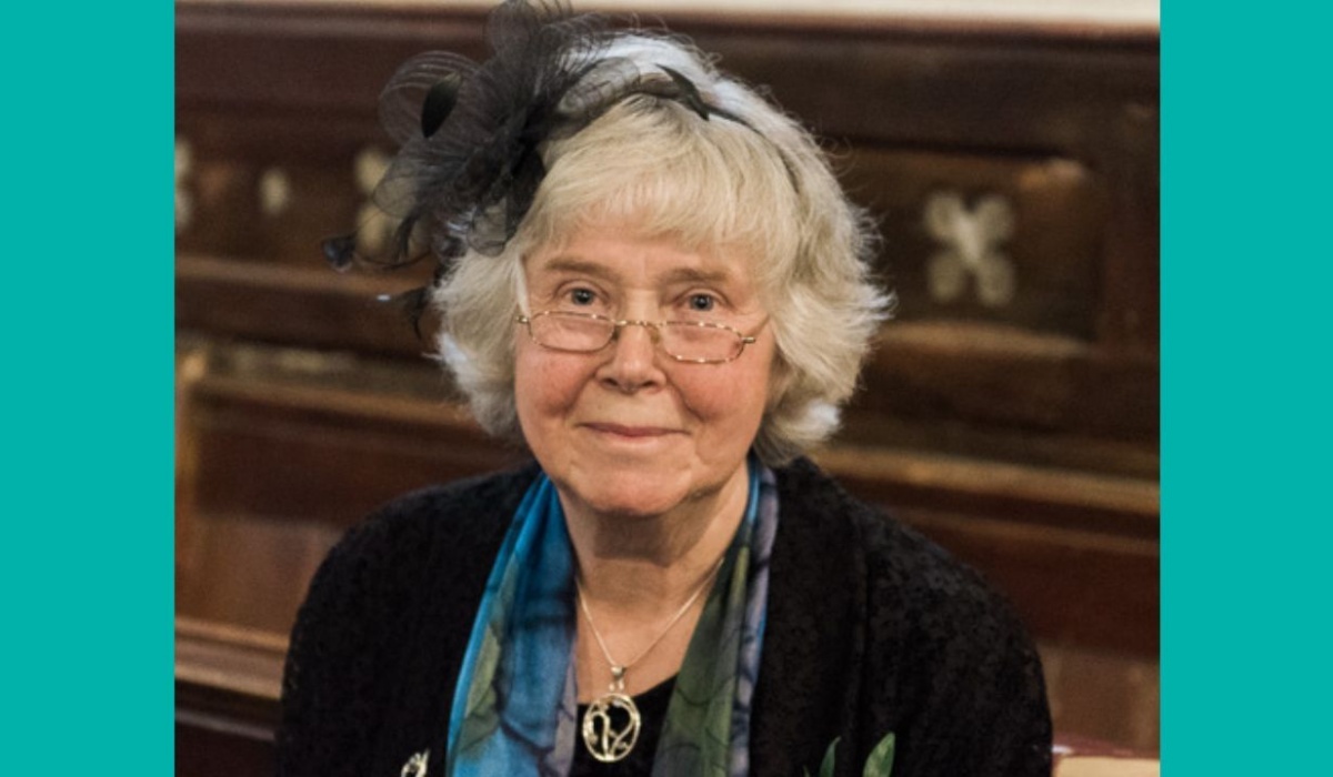A grey-haired woman dressed in blue for a wedding and wearing glasses, smiles at the camera from her pew.